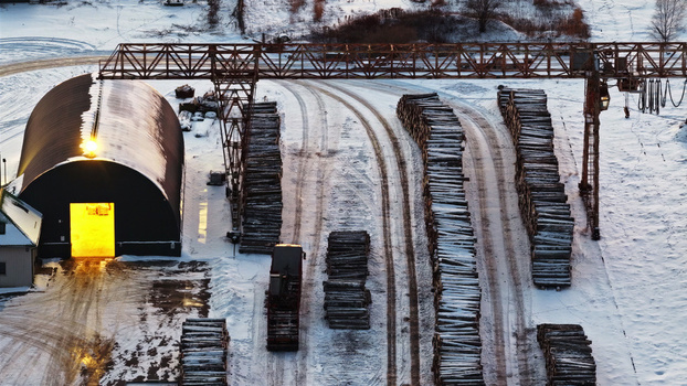 Aerial view of a snow covered lumber yard featuring stacks of logs a large gantry crane and a dark arched storage building with a glowing entrance in Latvia. Aerial view of a snow covered lumber yard featuring stacks of logs a large gantry crane and a dark arched storage building with a glowing entrance in Latvia.