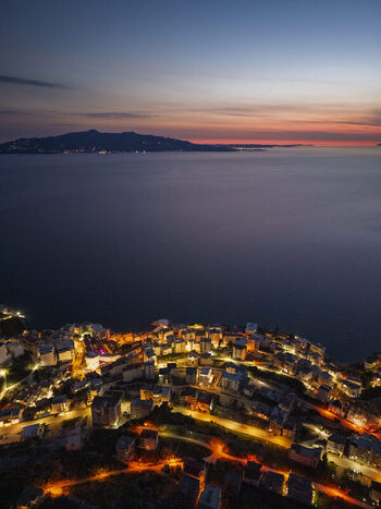 Aerial view of the coastal city of Saranda at twilight with illuminated buildings winding streets and the calm Ionian Sea under a colorful sunset sky in Saranda Vlore County Albania. Aerial view of the coastal city of Saranda at twilight with illuminated buildings winding streets and the calm Ionian Sea under a colorful sunset sky in Saranda Vlore County Albania.