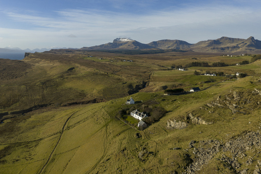 Aerial view of rugged green hills and white cottages nestled against the rocky slopes of the Trotternish Ridge under a clear blue sky Isle of Skye Scotland.