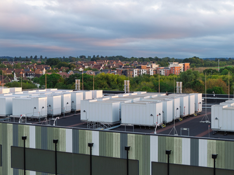 Aerial view of a data center rooftop featuring rows of white cooling units and infrastructure with residential buildings and trees in the background in the United Kingdom. Aerial view of a data center rooftop featuring rows of white cooling units and infrastructure with residential buildings and trees in the background in the United Kingdom.
