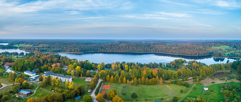 Aerial view of a serene lake surrounded by dense autumn forests small residential buildings and green fields under a clear blue sky in Latvia. Aerial view of a serene lake surrounded by dense autumn forests small residential buildings and green fields under a clear blue sky in Latvia.