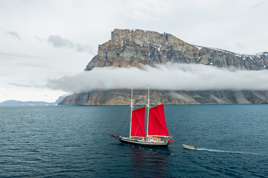 Aerial view of a sailboat with striking red sails navigating near the rocky cliffs of Storoen with low clouds clinging to the mountain slopes in Avannaata Kommunia Greenland. Aerial view of a sailboat with striking red sails navigating near the rocky cliffs of Storoen with low clouds clinging to the mountain slopes in Avannaata Kommunia Greenland.
