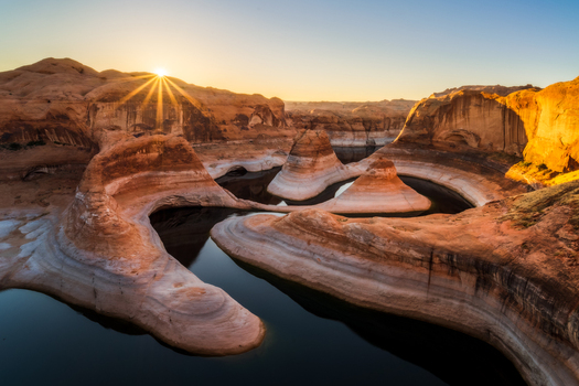 View of the winding sandstone formations of Reflection Canyon at sunrise with a sunburst over the desert landscape in Kane County Utah United States. View of the winding sandstone formations of Reflection Canyon at sunrise with a sunburst over the desert landscape in Kane County Utah United States.