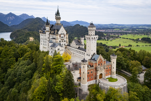 Aerial view of Neuschwanstein Castle perched atop a verdant hill its pale stone walls contrasting with the dark green forest below framed by distant mountains Neuschwanstein Casle Bayern Germany. Aerial view of Neuschwanstein Castle perched atop a verdant hill its pale stone walls contrasting with the dark green forest below framed by distant mountains Neuschwanstein Casle Bayern Germany.