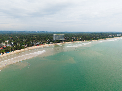 Aerial view of the sandy coastline and turquoise ocean with surfers in the water and a modern hotel building surrounded by palm trees in Weligama Southern Province Sri Lanka.