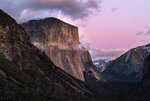 View of the granite monolith of El Capitan in Yosemite National Park with low hanging clouds and a purple sunset sky over the forest in the United States of America. View of the granite monolith of El Capitan in Yosemite National Park with low hanging clouds and a purple sunset sky over the forest in the United States of America.