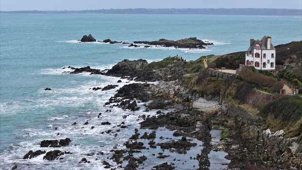 Aerial view of the rugged coastline of Locquirec featuring a white house with red shutters on a cliff overlooking turquoise waters and crashing waves Locquirec Brittany France.