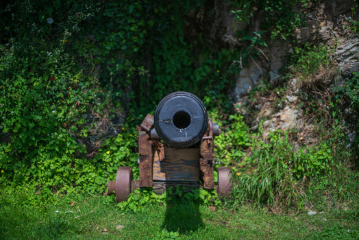 View of an antique iron cannon mounted on a weathered wooden carriage positioned against a lush foliage covered rocky bank in Charlestown Cornwall United Kingdom. View of an antique iron cannon mounted on a weathered wooden carriage positioned against a lush foliage covered rocky bank in Charlestown Cornwall United Kingdom.