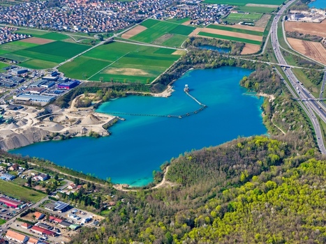 Aerial view of an aquamarine lake contrasting with the surrounding industrial area and lush greenery as a highway snakes nearby Sausheim Grand Est France.
