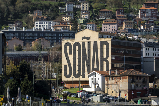 View of the large SONAR mural on a building facade with residential houses on a green hillside in the background under bright sunlight in Greater Bilbao Basque Country Spain. View of the large SONAR mural on a building facade with residential houses on a green hillside in the background under bright sunlight in Greater Bilbao Basque Country Spain.
