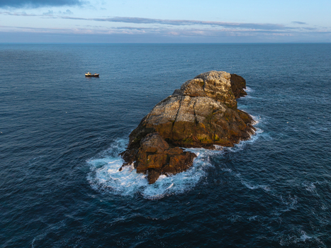 Aerial view of Sule Stack surrounded by deep blue ocean waters and crashing waves with a small boat nearby in Scotland United Kingdom. Aerial view of Sule Stack surrounded by deep blue ocean waters and crashing waves with a small boat nearby in Scotland United Kingdom.