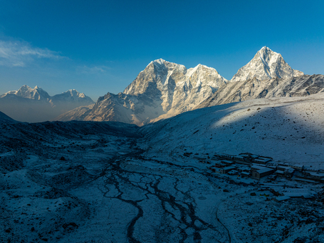 Aerial view of the snow covered Himalayan mountain range with a glacial valley and a small remote settlement nestled at the base under a clear blue sky in Nepal.