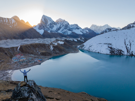 Aerial view of a person standing on a rocky outcrop overlooking the turquoise waters of the Gokyo Lakes surrounded by snow capped Himalayan peaks in Nepal.
