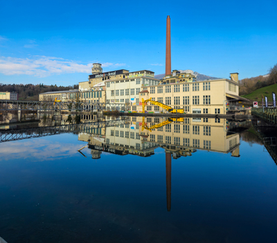 View of the Former Cellulose Factory with its tall chimney and yellow crane reflected in the calm river under a clear blue sky in Solothurn Solothurn Switzerland. View of the Former Cellulose Factory with its tall chimney and yellow crane reflected in the calm river under a clear blue sky in Solothurn Solothurn Switzerland.