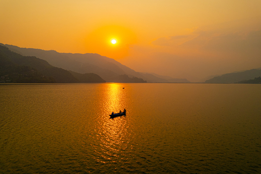 Aerial view of a small boat with people on a calm lake at sunset with mountains in the background under a golden sky in Nepal.