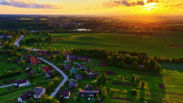 Aerial view of a rural village surrounded by lush green fields and trees under a vibrant golden sunset sky Latvia. Aerial view of a rural village surrounded by lush green fields and trees under a vibrant golden sunset sky Latvia.