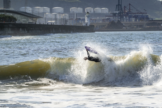 View of a bodyboarder performing a maneuver on a breaking wave at Ereaga Beach with the industrial harbor and lighthouse in the background in Algorta Euskadi Spain.
