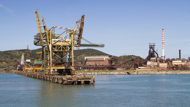 View of a large industrial port with cranes reflected in the still turquoise water backed by hills and a clear sky Piombino Tuscany Italy. View of a large industrial port with cranes reflected in the still turquoise water backed by hills and a clear sky Piombino Tuscany Italy.
