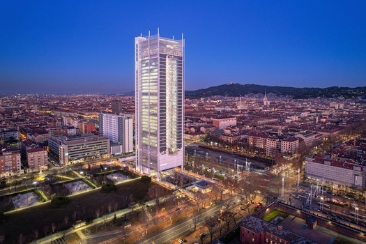 Aerial view of the Intesa Sanpaolo Skyscraper illuminated at dusk surrounded by the urban skyline city streets and distant hills under a deep blue sky Torino Piemonte Italy. Aerial view of the Intesa Sanpaolo Skyscraper illuminated at dusk surrounded by the urban skyline city streets and distant hills under a deep blue sky Torino Piemonte Italy.