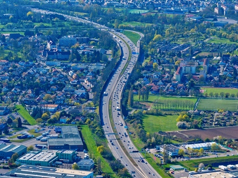 Aerial view of a serpentine highway cutting through urban sprawl bordered by vibrant green fields and dense trees Sausheim Grand Est France.