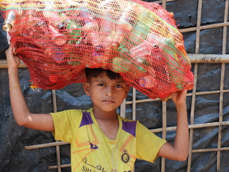 Cox s Bazar Chittagong Division Bangladesh 19 July 2026 View of a boy carrying a red mesh bag of plastic bottles in front of a bamboo shelter at a refugee camp. Cox s Bazar Chittagong Division Bangladesh 19 July 2026 View of a boy carrying a red mesh bag of plastic bottles in front of a bamboo shelter at a refugee camp.