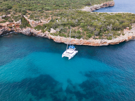 Aerial view of a white catamaran anchored in the turquoise waters of Cala Sa Nau cove surrounded by rocky cliffs and green trees in Felanitx Balearic Islands Spain. Aerial view of a white catamaran anchored in the turquoise waters of Cala Sa Nau cove surrounded by rocky cliffs and green trees in Felanitx Balearic Islands Spain.