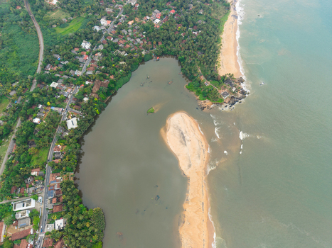 Aerial view of the coastline where a river meets the ocean with a sandbar lush tropical palm trees and residential houses along the road in Galle Southern Province Sri Lanka. Aerial view of the coastline where a river meets the ocean with a sandbar lush tropical palm trees and residential houses along the road in Galle Southern Province Sri Lanka.