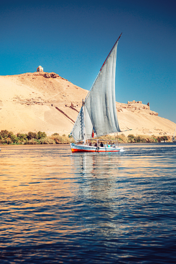 View of a traditional felucca sailboat with a large white sail on the Nile River near desert hills and ancient tombs under a clear blue sky in Aswan Aswan Governorate Egypt. View of a traditional felucca sailboat with a large white sail on the Nile River near desert hills and ancient tombs under a clear blue sky in Aswan Aswan Governorate Egypt.