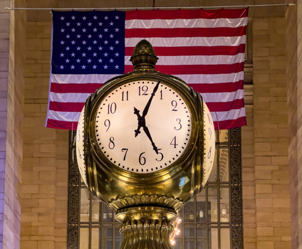 View of the iconic four faced opal clock in the main concourse of Grand Central Terminal with an American flag hanging in the background in New York New York United States. View of the iconic four faced opal clock in the main concourse of Grand Central Terminal with an American flag hanging in the background in New York New York United States.