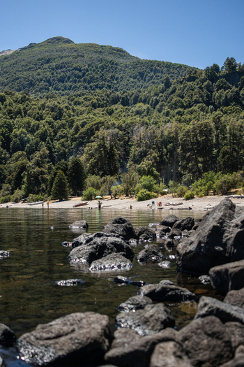 View of the rocky shoreline of Lago Paimun with clear water dense forest and mountains in the background in Huiliches Department Neuquen Province Argentina.