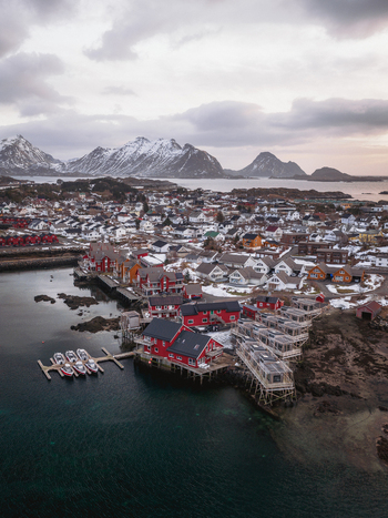 Aerial view of red and yellow houses nestled along the rugged coastline contrasting with the snow dusted mountains in the distance Ballstad Nordland Norway. Aerial view of red and yellow houses nestled along the rugged coastline contrasting with the snow dusted mountains in the distance Ballstad Nordland Norway.