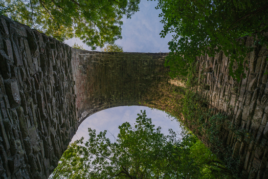View of the stone arch of the historic viaduct in Bodmin surrounded by lush green trees and foliage under a bright sky in Bodmin Cornwall United Kingdom. View of the stone arch of the historic viaduct in Bodmin surrounded by lush green trees and foliage under a bright sky in Bodmin Cornwall United Kingdom.