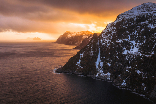 Aerial view of jagged snow dusted peaks plunging into the slate blue sea under a fiery sunset sky Nordland Nordland Norway.