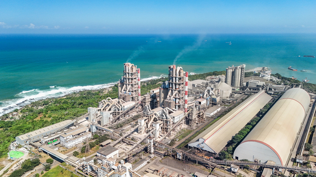 Aerial view of a large industrial cement factory complex with tall smokestacks storage domes and conveyor belts situated along the coastline in Indonesia.