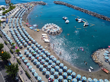 Aerial view of a crowded beach with blue and white umbrellas swimmers in the clear sea and stone breakwaters in San Lorenzo Liguria Italy.