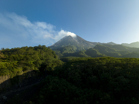 Aerial view of Mount Merapi with smoke rising from the crater above lush green forests and hills under a clear blue sky in Kecamatan Pakem Daerah Istimewa Yogya Indonesia. Aerial view of Mount Merapi with smoke rising from the crater above lush green forests and hills under a clear blue sky in Kecamatan Pakem Daerah Istimewa Yogya Indonesia.