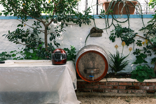 View of a wooden wine barrel and a red motorcycle helmet against a white wall with lush green plants in Qeparo Fshat Vlore County Albania. View of a wooden wine barrel and a red motorcycle helmet against a white wall with lush green plants in Qeparo Fshat Vlore County Albania.