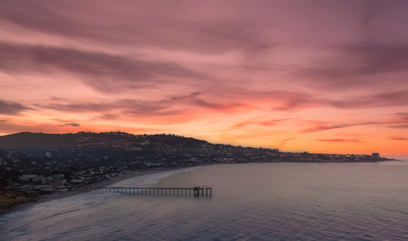 Aerial view of Scripps Pier extending into the Pacific Ocean at sunset with a vibrant pink and orange sky over the coastline of La Jolla California USA.