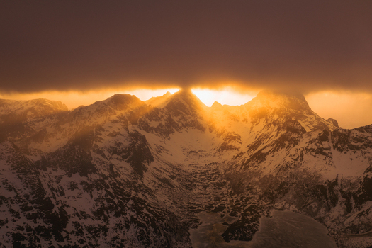 Aerial view of sun rays piercing through the clouds illuminating the snow capped peaks with a golden glow creating a dramatic contrast against the dark shadowed valleys Nordland Norway.