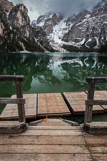 View of a wooden pier leading to a calm alpine lake with snow capped Dolomites mountains reflected in the water under a cloudy sky Trentino Alto Adige Italy. View of a wooden pier leading to a calm alpine lake with snow capped Dolomites mountains reflected in the water under a cloudy sky Trentino Alto Adige Italy.