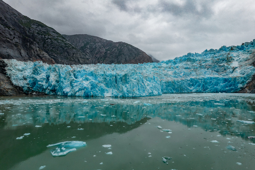 Aerial view of the Mendenhall Glacier with its bright blue ice face floating icebergs in the water and rugged mountains under a cloudy sky in Juneau Alaska United States. Aerial view of the Mendenhall Glacier with its bright blue ice face floating icebergs in the water and rugged mountains under a cloudy sky in Juneau Alaska United States.