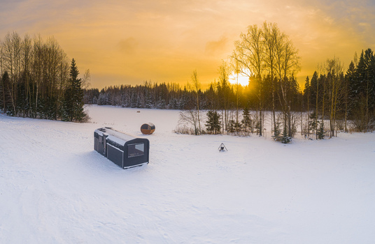 Aerial view of a modern modular cabin and a small round sauna pod in a snow covered landscape with a fire pit and forest under a golden sunset in Latvia. Aerial view of a modern modular cabin and a small round sauna pod in a snow covered landscape with a fire pit and forest under a golden sunset in Latvia.