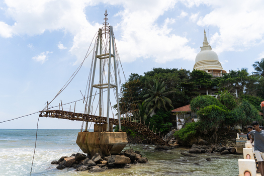 View of the Parevi Duwa Temple featuring a white stupa and a metal bridge structure over the rocky coast and ocean waves Matara Southern Province Sri Lanka.