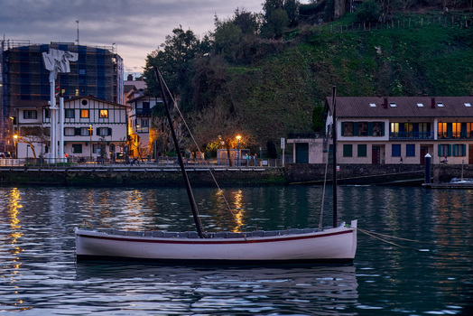 Pasai Donibane Spain 09 March 2026 View of a traditional wooden boat moored along the main harbor street with historic buildings and a lush green hillside at twilight.