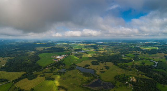 Aerial view of the lush green rural landscape featuring rolling fields dense forests small lakes and a winding road under a dramatic cloudy sky in Latvia. Aerial view of the lush green rural landscape featuring rolling fields dense forests small lakes and a winding road under a dramatic cloudy sky in Latvia.