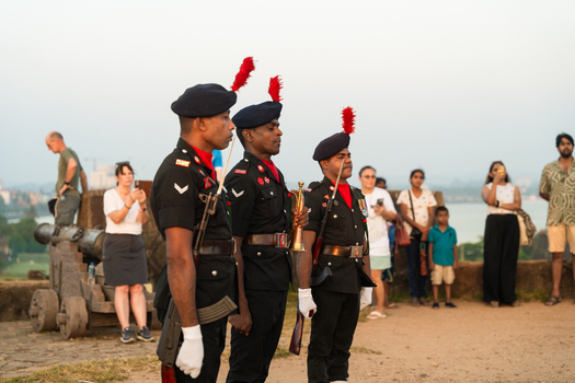 Sri Lanka 24 January 2026 View of three soldiers in ceremonial black uniforms with red plumes standing at attention near a historic cannon with tourists in the background.