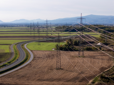 Aerial view of high voltage power lines and electricity pylons stretching across agricultural fields and a winding road with mountains in the distance Slovenia.