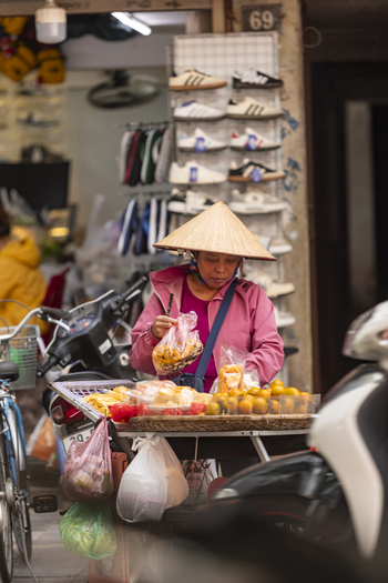 Hanoi Vietnam 11 November 2025 View of a street vendor wearing a conical hat selling fresh fruit from a cart on a busy street with motorbikes and a shoe shop in the background.