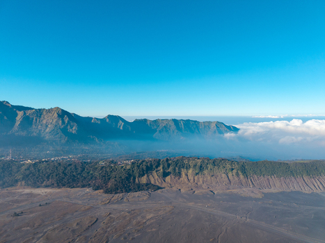 Aerial view of Mount Bromo landscape featuring rugged volcanic ridges vast sandy plains and low hanging clouds under a clear blue sky in Tosari East Java Indonesia.