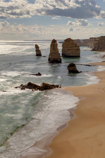 View of the Twelve Apostles limestone stacks rising from the ocean along a sandy beach with crashing waves and high cliffs under a cloudy sky in Australia. View of the Twelve Apostles limestone stacks rising from the ocean along a sandy beach with crashing waves and high cliffs under a cloudy sky in Australia.